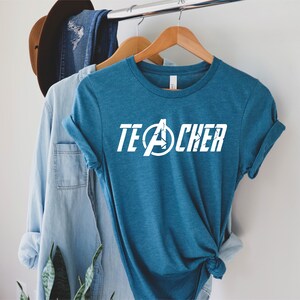 Op de afbeelding: Een tealblauw t-shirt met een witte afbeelding waarop "TEACHER" staat met een Avengers-logo in het midden van het woord.