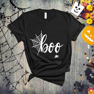 May include: Black t-shirt with a white spiderweb and the word "boo" in a white cursive font. A small white spider hangs from the bottom of the word "boo".