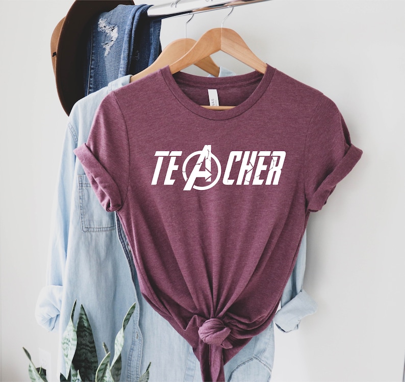 Op de afbeelding: Bordeauxkleurig T-shirt met een witte afbeelding van het Avengers-logo en het woord "TEACHER" erop gedrukt.