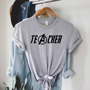 Op de afbeelding: Grijs T-shirt met een zwarte afbeelding van het Avengers-logo en het woord "TEACHER" erop gedrukt.
