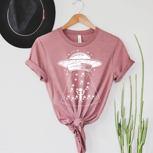 May include: A pink t-shirt with a white graphic of a UFO hovering over a field of marijuana plants. The graphic also includes a small alien figure with its arms raised.