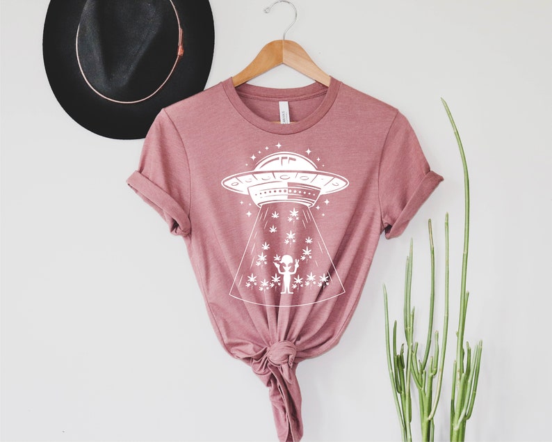 May include: A pink t-shirt with a white graphic of a UFO hovering over a field of marijuana plants. The graphic also includes a small alien figure with its arms raised.