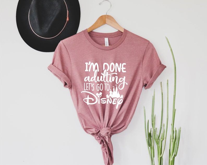 May include: A pink t-shirt with the text "I'm done adulting let's go to Disney" in white lettering. The shirt has a knot tied at the bottom.