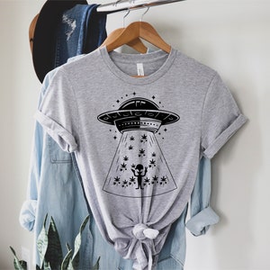 May include: A gray t-shirt with a black graphic of an alien in a flying saucer beaming down a marijuana plant. The graphic is surrounded by stars.