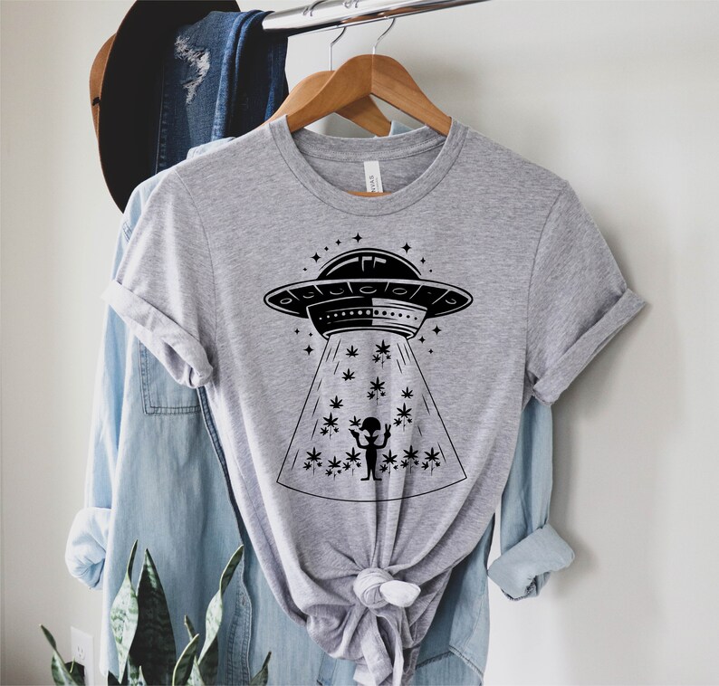 May include: A gray t-shirt with a black graphic of an alien in a flying saucer beaming down a marijuana plant. The graphic is surrounded by stars.