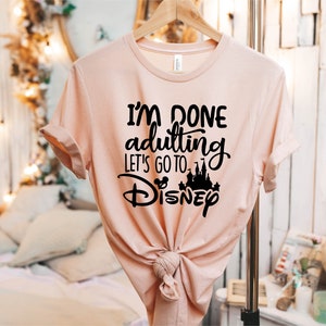 May include: A pink t-shirt with the text "I'm done adulting let's go to Disney" in black lettering. The text is in a handwritten style font. A black silhouette of Cinderella's castle is included in the text.