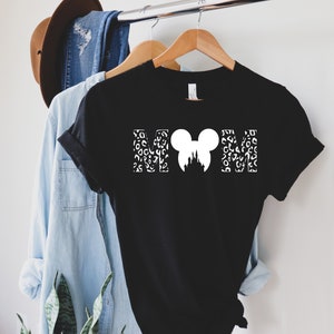 Puede incluir: Camiseta negra con un gr&aacute;fico blanco de las letras "MOM" con una cabeza de Mickey Mouse y una silueta del castillo de Cenicienta en el medio. Las letras est&aacute;n rellenas con un patr&oacute;n de leopardo blanco.