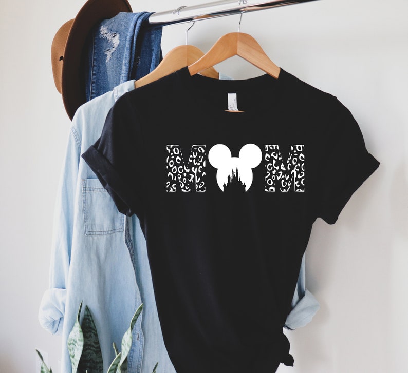 Puede incluir: Camiseta negra con un gr&aacute;fico blanco de las letras "MOM" con una cabeza de Mickey Mouse y una silueta del castillo de Cenicienta en el medio. Las letras est&aacute;n rellenas con un patr&oacute;n de leopardo blanco.