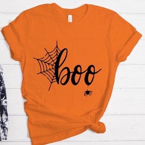 May include: Orange t-shirt with a black spider web and the word "boo" in a handwritten font. A small black spider hangs from the bottom of the word "boo".