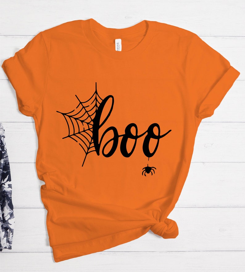 May include: Orange t-shirt with a black spider web and the word "boo" in a handwritten font. A small black spider hangs from the bottom of the word "boo".