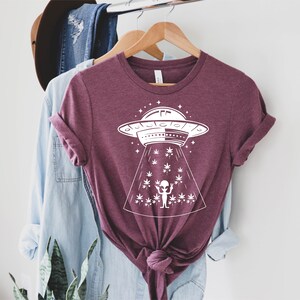 May include: A maroon t-shirt with a white graphic of an alien in a flying saucer above a field of marijuana plants.