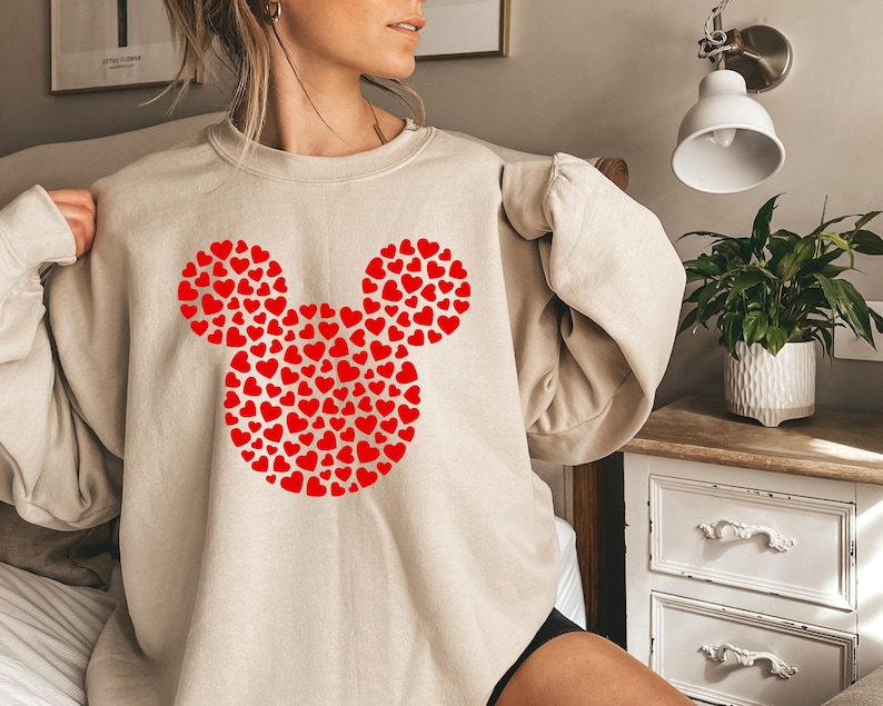 Puede incluir: Una sudadera beige con un dise&ntilde;o de Mickey Mouse en forma de coraz&oacute;n rojo.