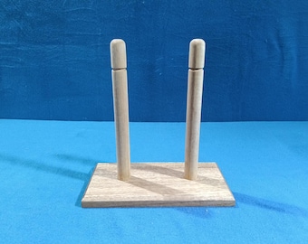 May include: A wooden display stand with two vertical dowels and a rectangular base. The stand is made of light-colored wood and is designed to hold an item upright. The background is a solid blue.