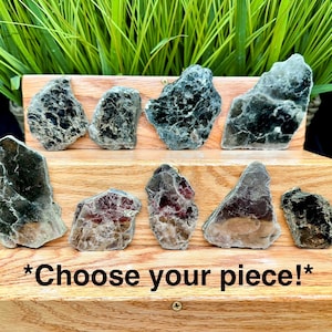 May include: A collection of raw mica stone pieces in various shapes and sizes, presented on a wooden stand. The stones display a range of colours, including black, grey, and reddish-brown. The text "Choose your piece!" is visible.