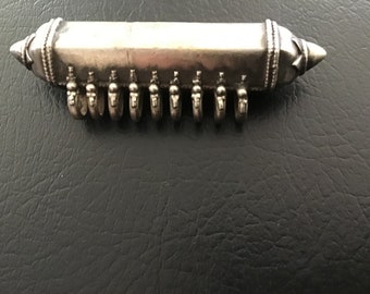 May include: A silver-colored, oblong pendant with a cylindrical body and conical ends. The pendant has a series of small loops along the bottom edge. The piece is likely jewelry.