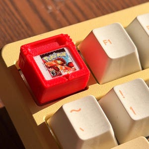 Puede incluir: Una tecla roja, impresa en 3D, con forma de cartucho de videojuego retro, con una etiqueta de juego colorida. La tecla está colocada en un teclado amarillo con teclas blanquecinas, incluida una tecla F1.