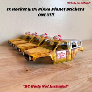 May include: A row of yellow toy car bodies with Pizza Planet decals, each topped with a rocket and pizza-themed decoration. The text "1x Rocket & 2x Pizza Planet Stickers ONLY!!!" is visible. "RC Body Not Included" is also displayed.