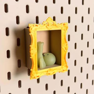 May include: A bright yellow, ornate, rectangular picture frame mounted on a pegboard. Inside the frame, a small, pale, translucent, smooth, rounded figure with two visible legs is positioned in a seated, reclining posture against the back wall.