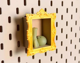 May include: A bright yellow, ornate, rectangular picture frame mounted on a pegboard. Inside the frame, a small, pale, translucent, smooth, rounded figure with two visible legs is positioned in a seated, reclining posture against the back wall.