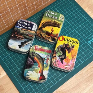 May include: Four rectangular metal tins featuring vintage magazine cover illustrations of fishing scenes. The tins display the titles Field & Stream, Outdoorsman, and Outdoor Life, with colorful graphics of fish, anglers, and natural landscapes.