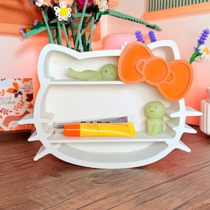 May include: A white wall-mounted shelf shaped like a cat head with a large orange bow on the right side. The shelf has two horizontal tiers holding two small pale green figurines and two tubes of skincare products with orange and silver caps.