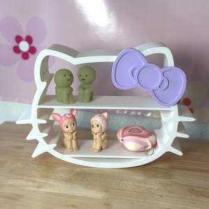 Puede incluir: Una estantería blanca de dos niveles con forma de cabeza de gato y un lazo morado. La estantería contiene pequeñas figuras, incluyendo dos figuras verdes, dos figuras con sombreros y un objeto a rayas rosas y blancas.