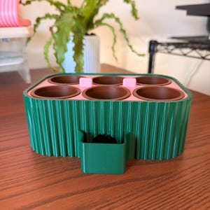 May include: A green and pink planter with six brown pots. The planter has a ribbed exterior and a small attached container. The pots are arranged in two rows of three. The planter is sitting on a wooden surface.