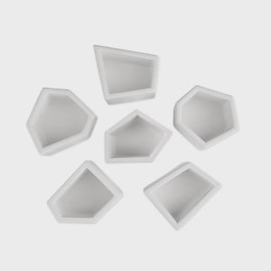May include: Six white, geometric-shaped containers arranged on a light gray background. Each container has a pentagonal or irregular shape with a recessed interior. The containers are empty and appear to be made of a smooth material.