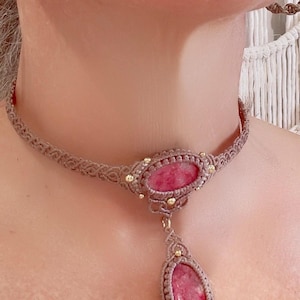 May include: A handmade macrame necklace featuring a pink gemstone pendant. The necklace is a light brown color with gold accents. The pendant is an oval shape, and the necklace is worn around the neck.