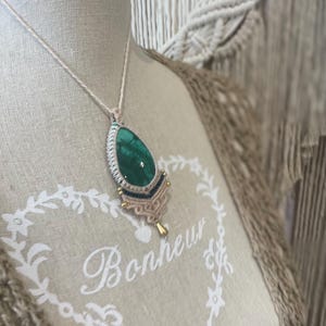 May include: A handcrafted macrame necklace featuring a teardrop-shaped, vibrant green malachite stone pendant. The pendant is accented with gold-colored beads and a delicate cream-colored cord. The necklace is displayed on a mannequin.