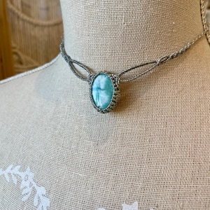 Op de afbeelding: Een handgemaakte macramé ketting met een lichtblauwe ovale edelsteen hanger. De hanger is gezet in een zilverkleurig macramé ontwerp en de ketting heeft een delicate, geweven koord. De ketting wordt op een mannequin getoond.