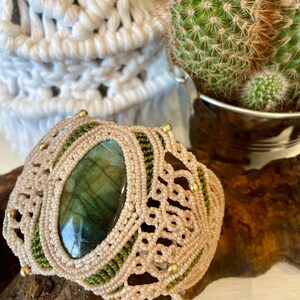 May include: A handmade macrame bracelet featuring a large, oval labradorite stone. The bracelet is beige with green accents and small gold-colored beads. The stone has shades of green and blue. The bracelet is displayed on a piece of wood.