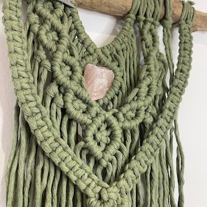 May include: An olive green macrame wall hanging, with a natural wooden dowel. A rose quartz crystal is set within the knotted design. The tag reads "Hand Made".
