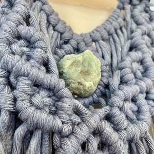 May include: A close-up shot of a handmade macrame wall hanging in a muted blue hue. The woven design incorporates a light green stone. The macrame is knotted in a decorative pattern, showcasing the craftmanship.