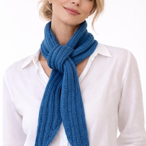 May include: A vibrant blue knitted scarf, knotted at the neck. The scarf is made of a ribbed knit fabric and features a small brown leather tag. The scarf is worn over a white button-down shirt.
