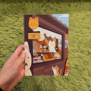 Könnte beinhalten: Illustration einer Katzenbäckerei mit brauner Holzfassade. Das Schaufenster zeigt Eiswaffeln, Gebäck und Brot. Ein Schild mit japanischem Text und einem Katzenkopf hängt darüber. Eine Katze greift nach dem Fenster.