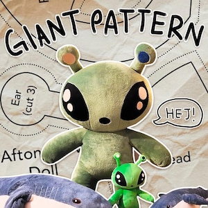 Puede incluir: Un muñeco de peluche verde alienígena con grandes ojos negros y pequeñas antenas. El muñeco tiene una expresión amigable y está rodeado por un muñeco alienígena verde más pequeño y un patrón con el texto "GIANT PATTERN" y "HEJ!".