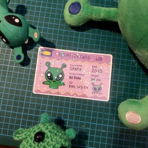 Op de afbeelding: Een lila alien-identiteitskaart met de naam SPARV en AFTON, geboren in 2023, en een lengte van 34 cm. De kaart toont een cartoon alien illustratie en de ID 805.515.59. De kaart is omgeven door groene alien knuffels.