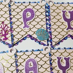 Pu&ograve; includere: Uno striscione a tema sirenetta con lettere di glitter viola che compongono la parola "Happy" e una conchiglia di glitter blu. Lo striscione &egrave; decorato con un motivo a squame di sirena rosa e oro.