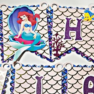 Pu&ograve; includere: Uno striscione di compleanno con una sirena su sfondo viola e blu. Lo striscione presenta una sirena, un cavalluccio marino e le lettere "Happy".
