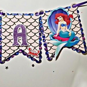 Pu&ograve; includere: Uno stendardo viola e dorato con una sirena seduta su una conchiglia. Lo stendardo ha un bordo a festoni e una lettera "A" di glitter viola. C'&egrave; un granchio rosso sullo stendardo.