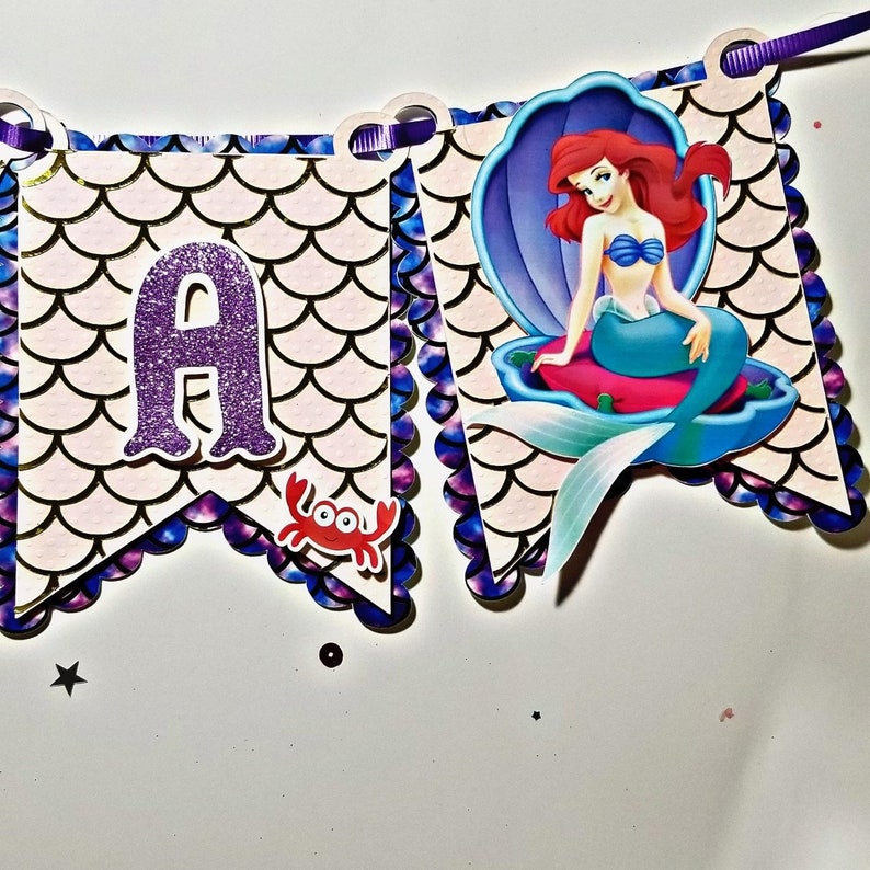 Pu&ograve; includere: Uno stendardo viola e dorato con una sirena seduta su una conchiglia. Lo stendardo ha un bordo a festoni e una lettera "A" di glitter viola. C'&egrave; un granchio rosso sullo stendardo.