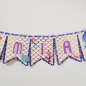 Pu&ograve; includere: Uno striscione viola e oro con il nome "MIA" scritto in lettere viola. Lo striscione ha un tema di sirena con immagini di Ariel de La Sirenetta e uno schema di conchiglie.