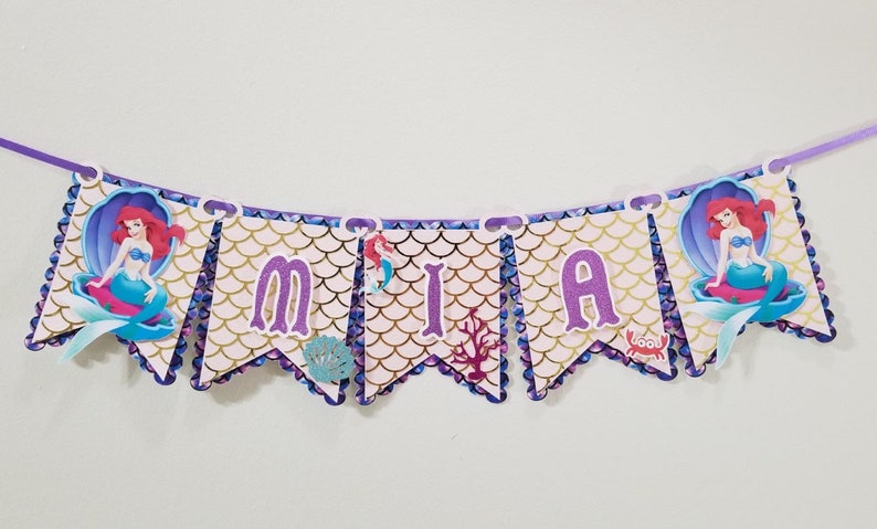 Pu&ograve; includere: Uno striscione viola e oro con il nome "MIA" scritto in lettere viola. Lo striscione ha un tema di sirena con immagini di Ariel de La Sirenetta e uno schema di conchiglie.