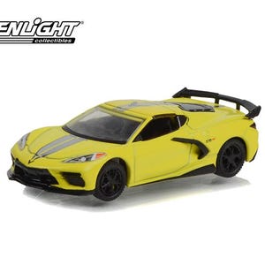 May include: A yellow die-cast model sports car with black wheels, a rear spoiler, and dual grey racing stripes running along the hood and roof. The Greenlight Collectibles logo is visible in the top left corner of the image.