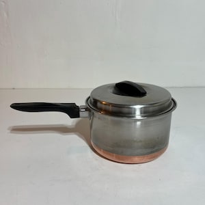 May include: A stainless steel saucepan with a copper bottom and a black handle. The pot has a lid with a black handle. The saucepan is designed for cooking and is suitable for various kitchen tasks.