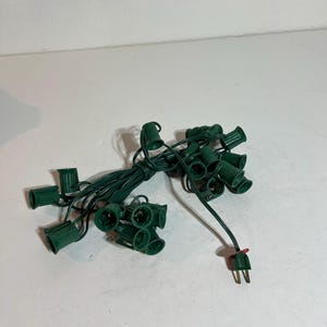 May include: A bundle of green Christmas light sockets and wiring, with a two-prong plug. The sockets are cylindrical and evenly spaced along the dark green wire. The plug has two prongs and a small red accent.