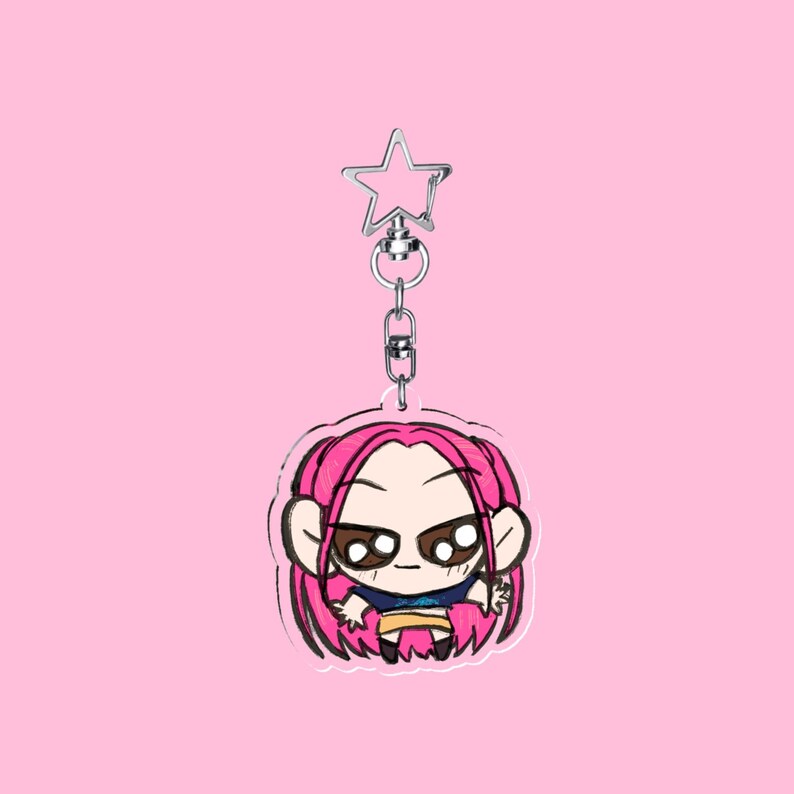 Puede incluir: Un llavero de dibujos animados con un personaje de pelo rosa, ojos grandes y una camisa azul. El llavero tiene un cierre plateado en forma de estrella y un fondo rosa.