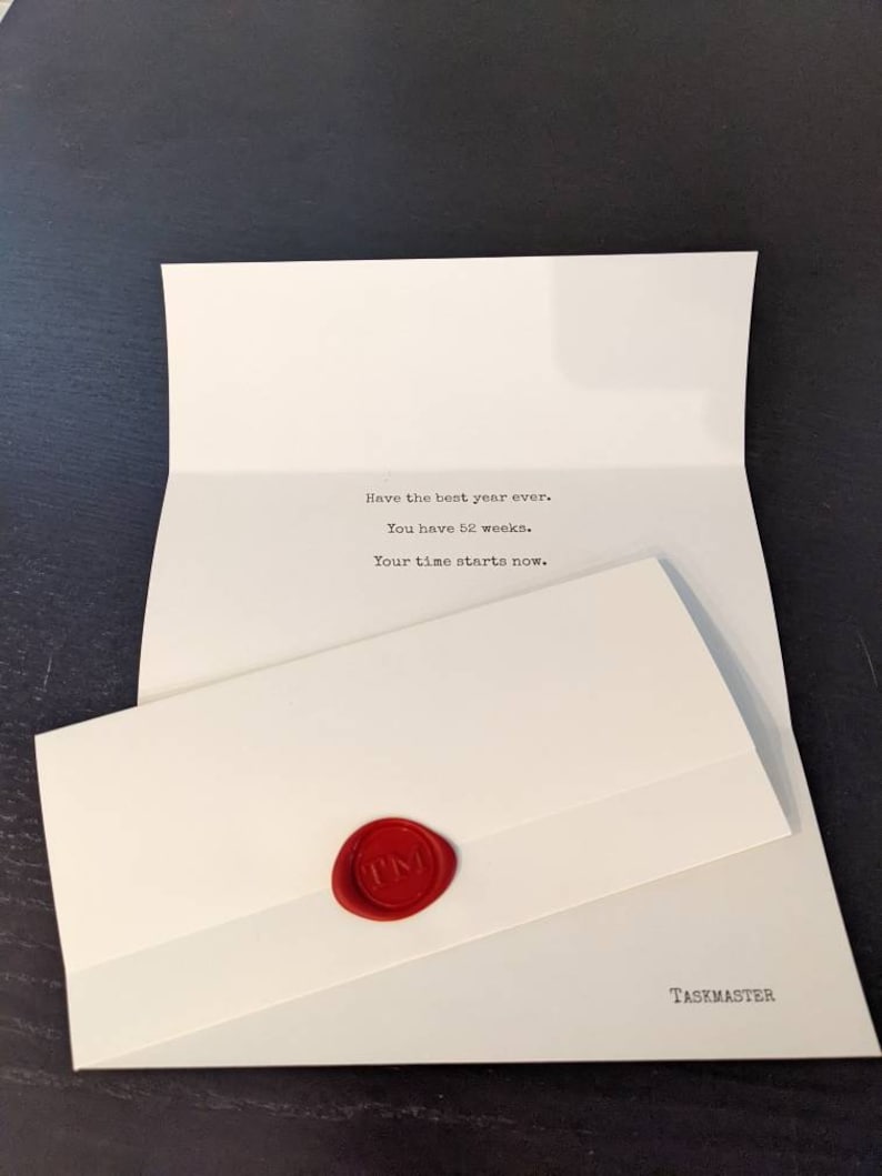 Puede incluir: Una tarjeta blanca abierta con el texto "Have the best year ever. You have 52 weeks. Your time starts now." y un sobre m&aacute;s peque&ntilde;o sellado con un sello de cera rojo. La palabra "TASKMASTER" est&aacute; impresa en la parte inferior de la tarjeta.