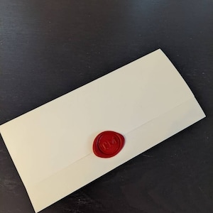 Puede incluir: Sobre de color marfil sellado con un sello de cera rojo. El sello presenta las iniciales "TM" en un dise&ntilde;o circular. El sobre est&aacute; colocado en diagonal sobre una superficie oscura.